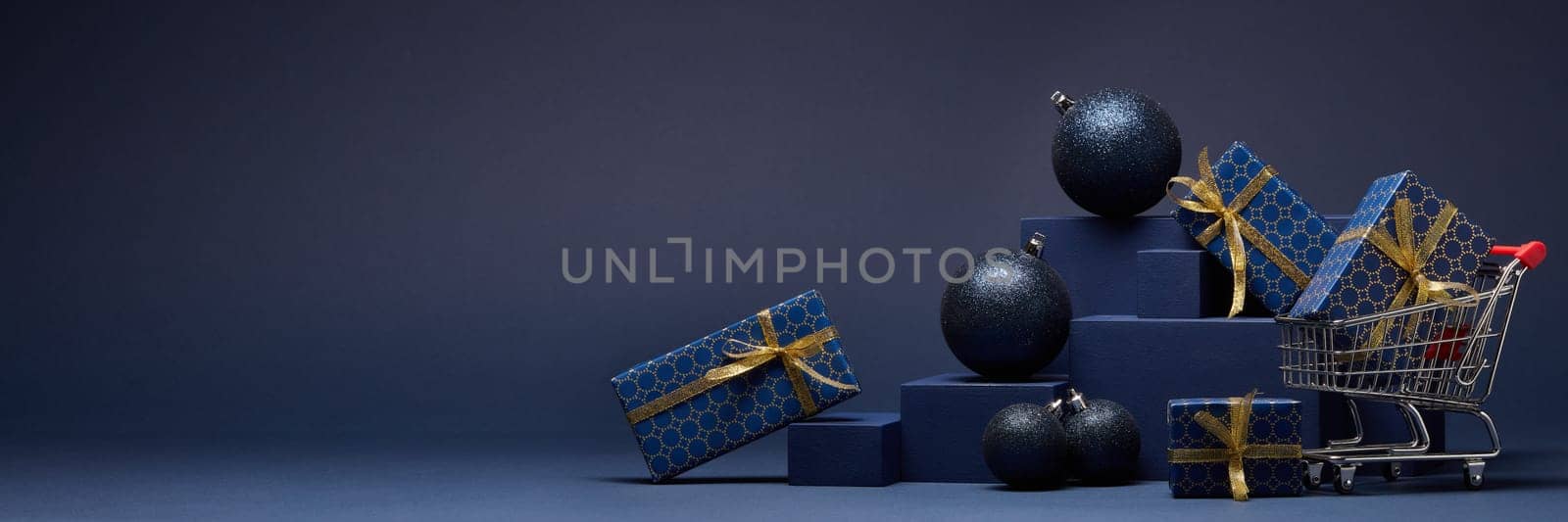 Shopping trolley, gift boxes and Christmas ornaments on dark blue background with copy space. Christmas and New Year shopping. Winter sale. For design of advertising, greeting card