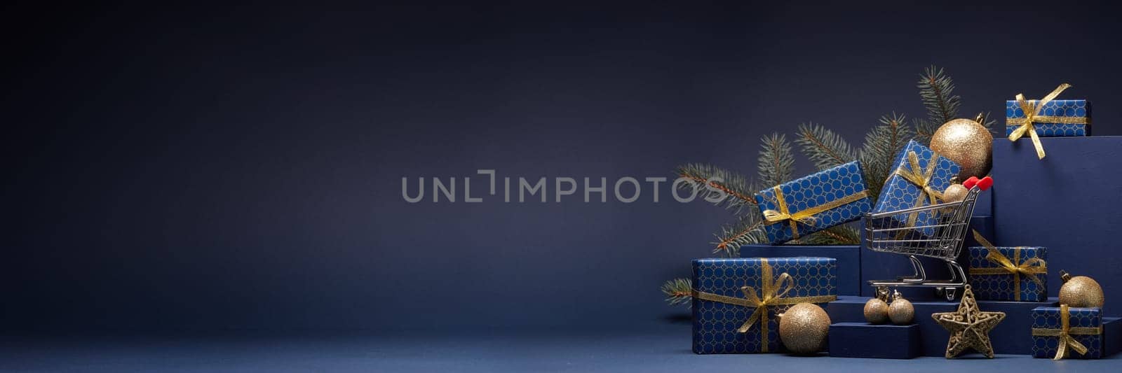 Shopping trolley, gift boxes, fir branch and Christmas ornaments on dark blue background with copy space. Christmas and New Year shopping. Winter sale. For design of advertising, greeting card