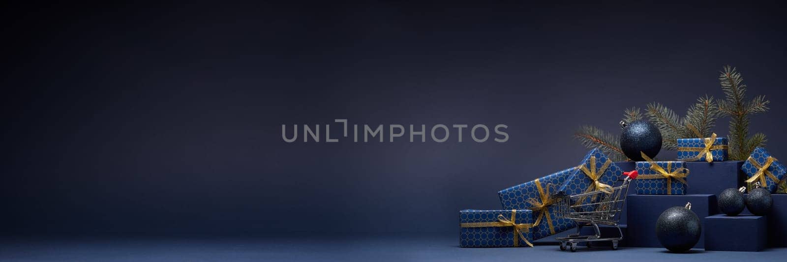 Shopping trolley, gift boxes, fir branch and Christmas ornaments on dark blue background with copy space. Christmas and New Year shopping. Winter sale. For design of advertising, greeting card