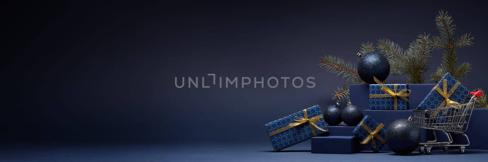 Shopping trolley, gift boxes, fir branch and Christmas ornaments on dark blue background with copy space. Christmas and New Year shopping. Winter sale. For design of advertising, greeting card