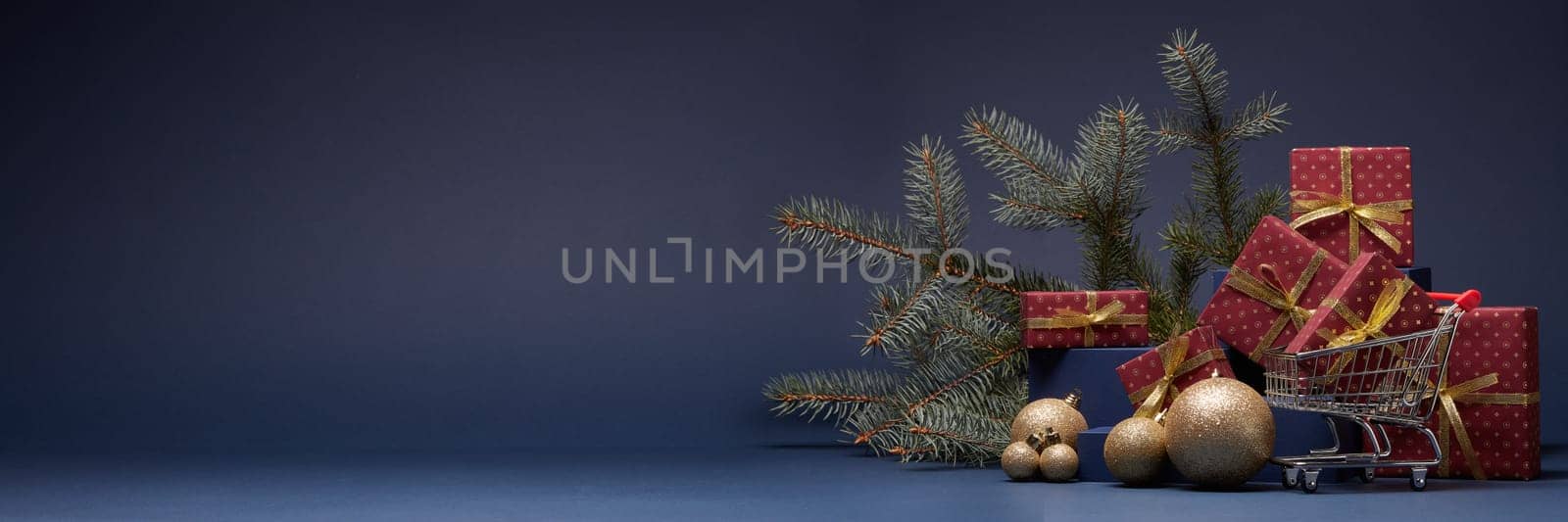 Shopping trolley, red gift boxes, fir branch and Christmas ornaments on dark blue background with copy space. Christmas and New Year shopping. Winter sale. For design of advertising, greeting card