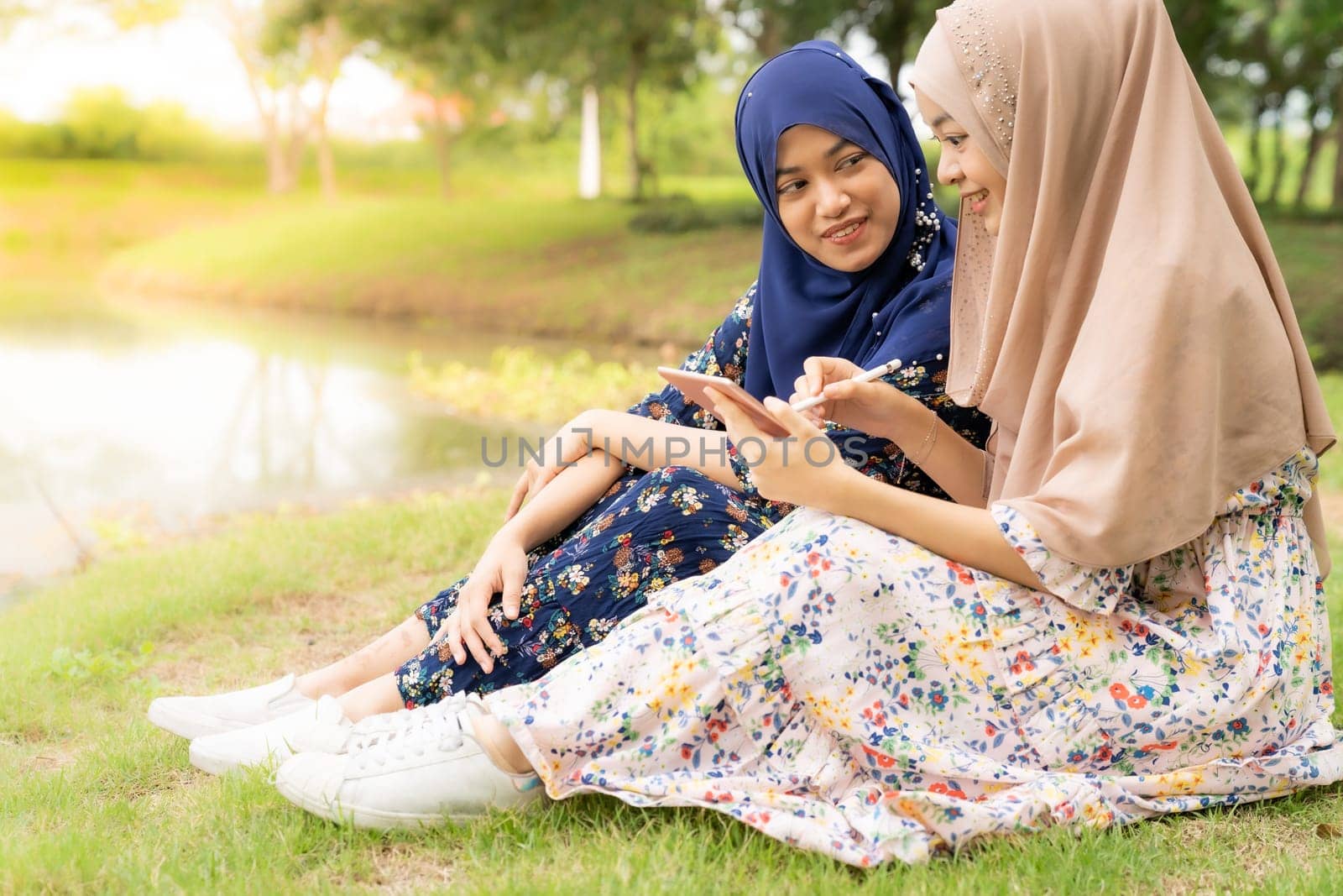 Young adult teenager Muslim Islamic Thai Asian women using smartphones and digital tablet in social media beside lake and green park in University college school.