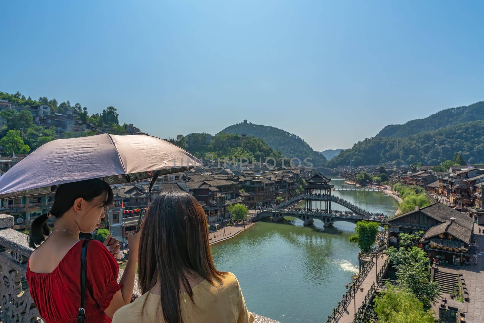 Feng Huang, China -  August 2019 : Women holding umbrella standing on the bridge and admiring the traditional old houses on the riverbanks of Tuo river in Feng huang Old Town
