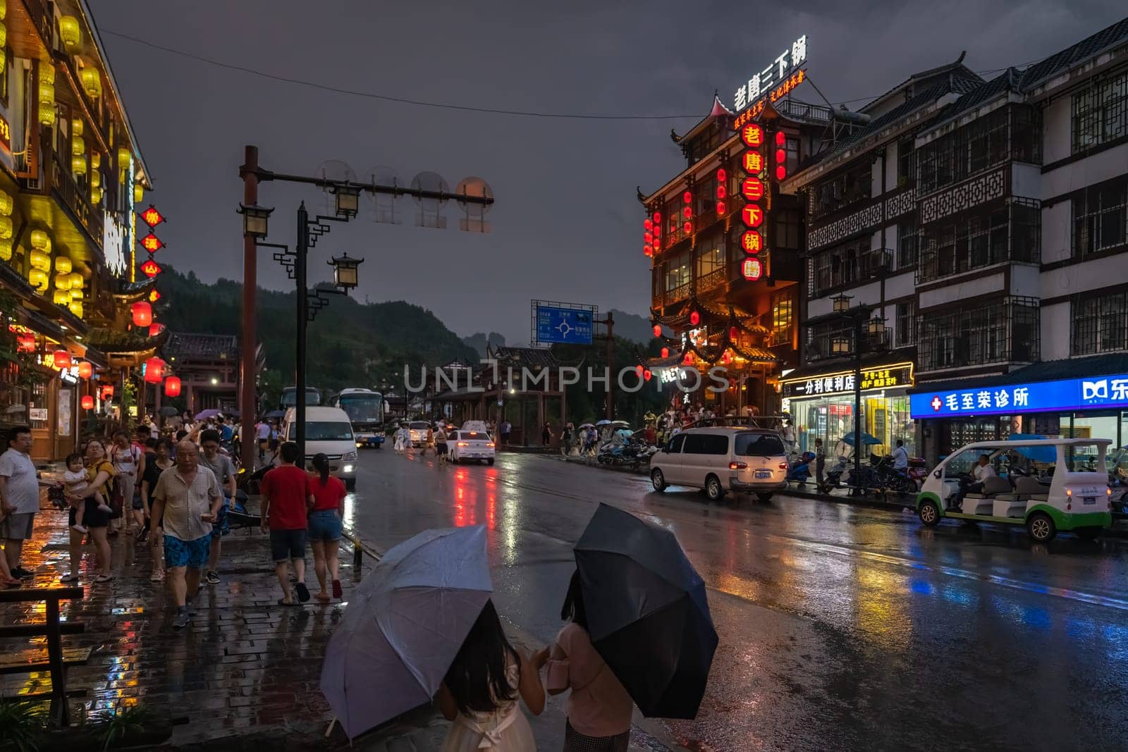Wulingyuan, China - August 2019 : People walking in the rain with lights on buildings lit at night at a busy bustling high street in Wulingyuan town at dusk