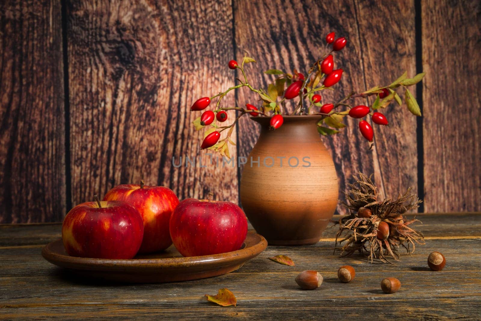 Autumnal arrangement with three red apples on a clay plate, rosehip branches in a clay vase and hazelnuts.