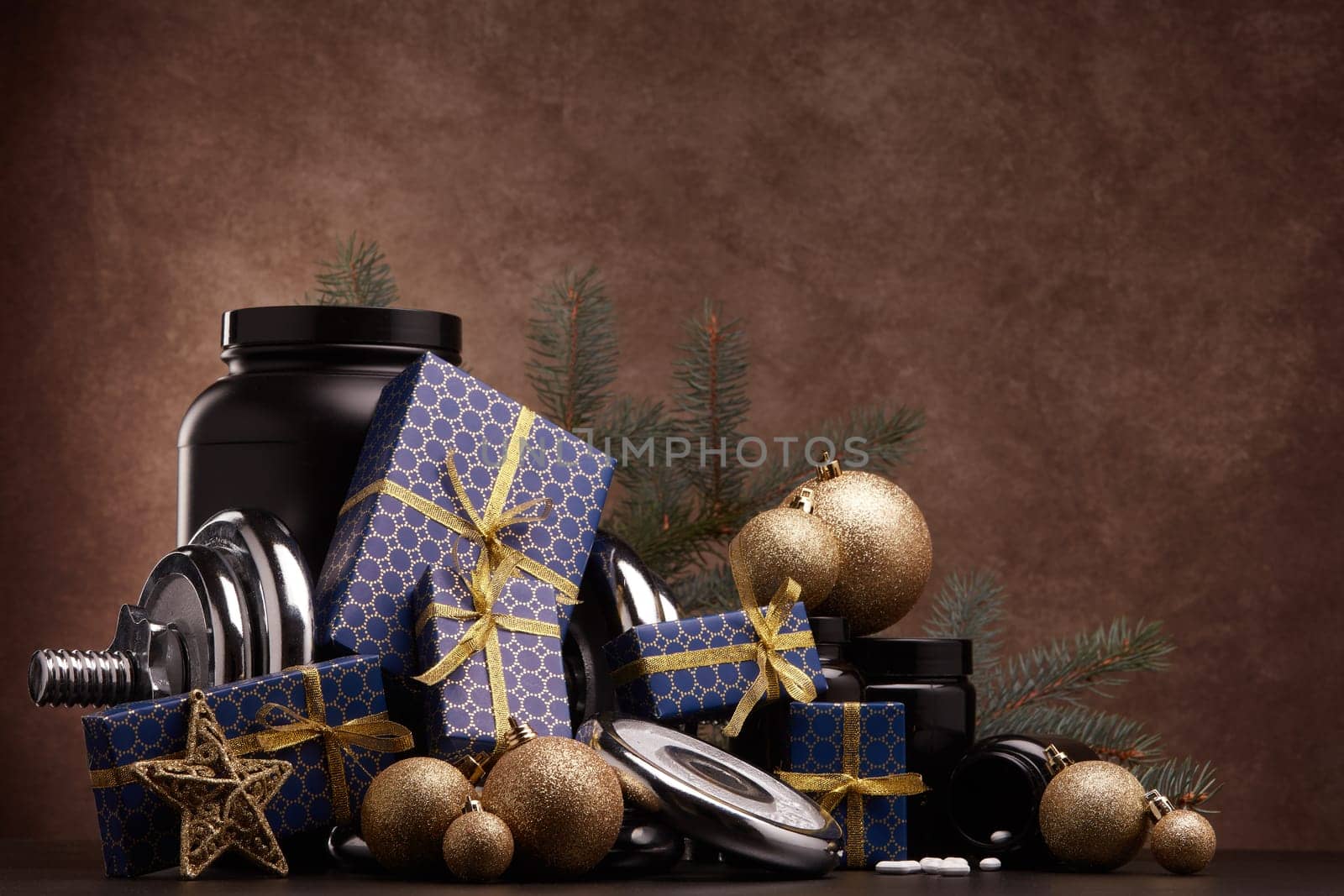 Sports nutrition (supplements), sports equipment, fir branch, gift boxes, Christmas ornaments on dark brown background with copy space. Fitness, sport, healthy lifestyle concept. New Year. Christmas.