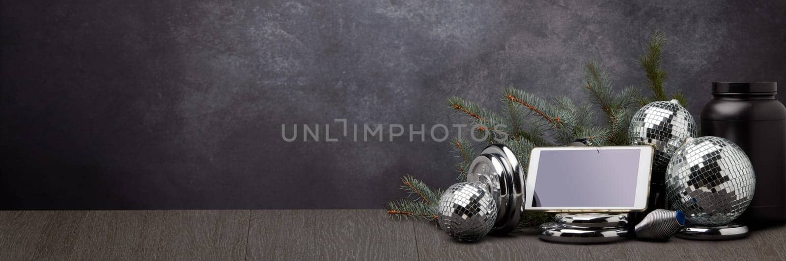 Sports nutrition, sports equipment, fir branch, Christmas ornament, blank computer tablet on dark grey background with copy space. Fitness, sport or healthy lifestyle concept. New Year and Christmas.