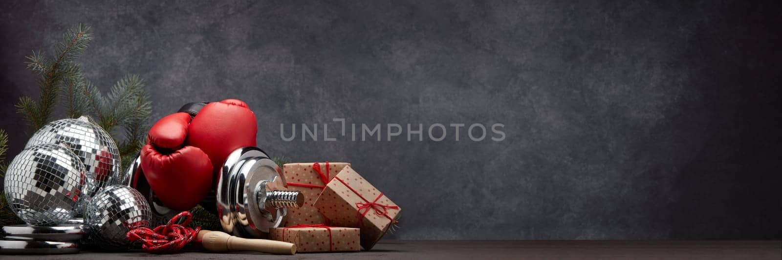 Sports equipment: dumbbell, boxing gloves, skipping rope, fir branch, Christmas ornament, gifts on dark grey background with copy space. Fitness, sport, healthy lifestyle concept. New Year. Christmas
