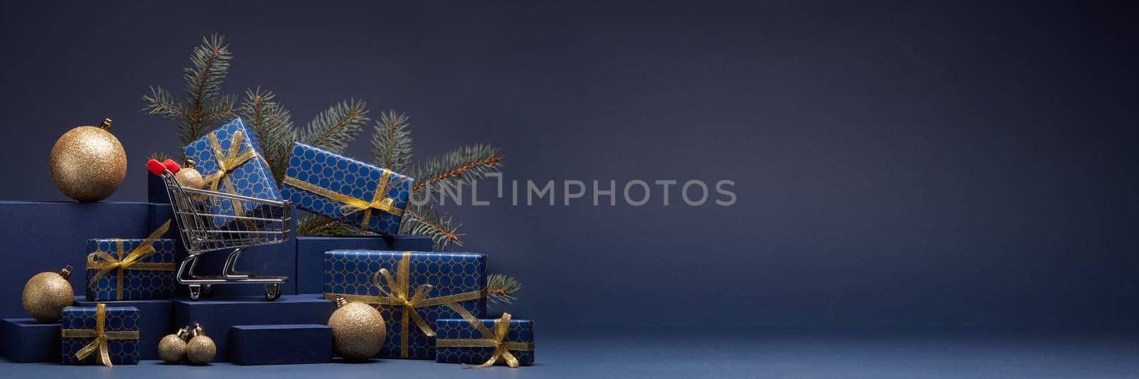 Shopping trolley, gift boxes, fir branch and Christmas ornaments on dark blue background with copy space. Christmas and New Year shopping. Winter sale. For design of advertising, greeting card