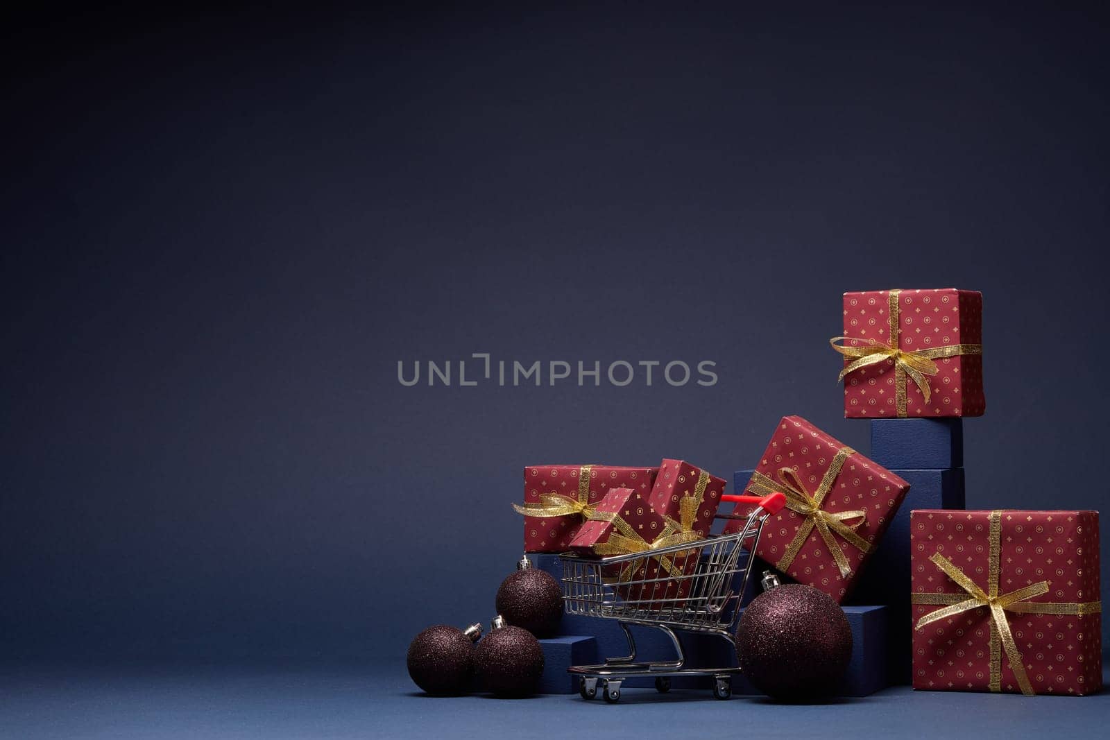 Shopping trolley, red gift boxes, fir branch and Christmas ornaments on dark blue background with copy space. Christmas and New Year shopping. Winter sale. For design of advertising, greeting card