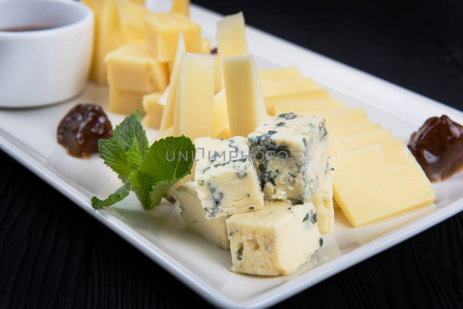 Cheese plate on a dark background table. Many kinds of cheese with sauce and greens on a white plate, closeup shot