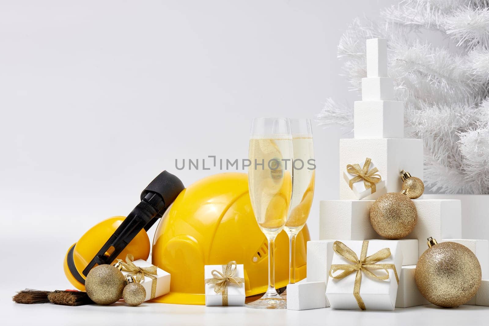 Construction protective earphones and hard hat, fir tree, flutes with champagne, gift boxes and Christmas ornament on a white background. New Year and Christmas construction