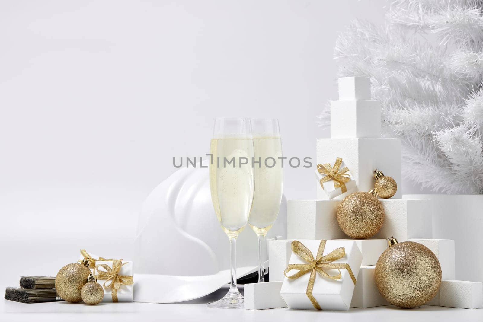 Construction hard hat, white fir tree, two glasses with champagne, gift boxes and Christmas ornaments on a white background. New Year and Christmas construction background