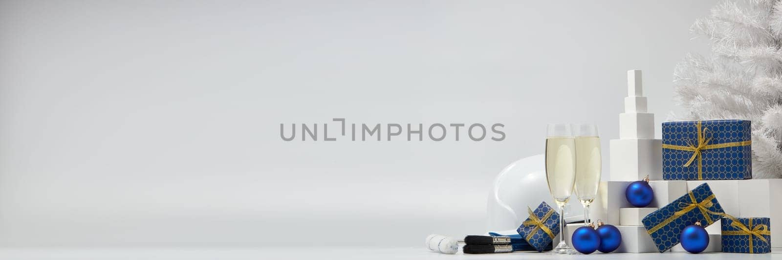 Construction hard hat, white fir tree, two glasses with champagne, gift boxes and Christmas ornaments on a white background with copy space. New Year and Christmas construction background