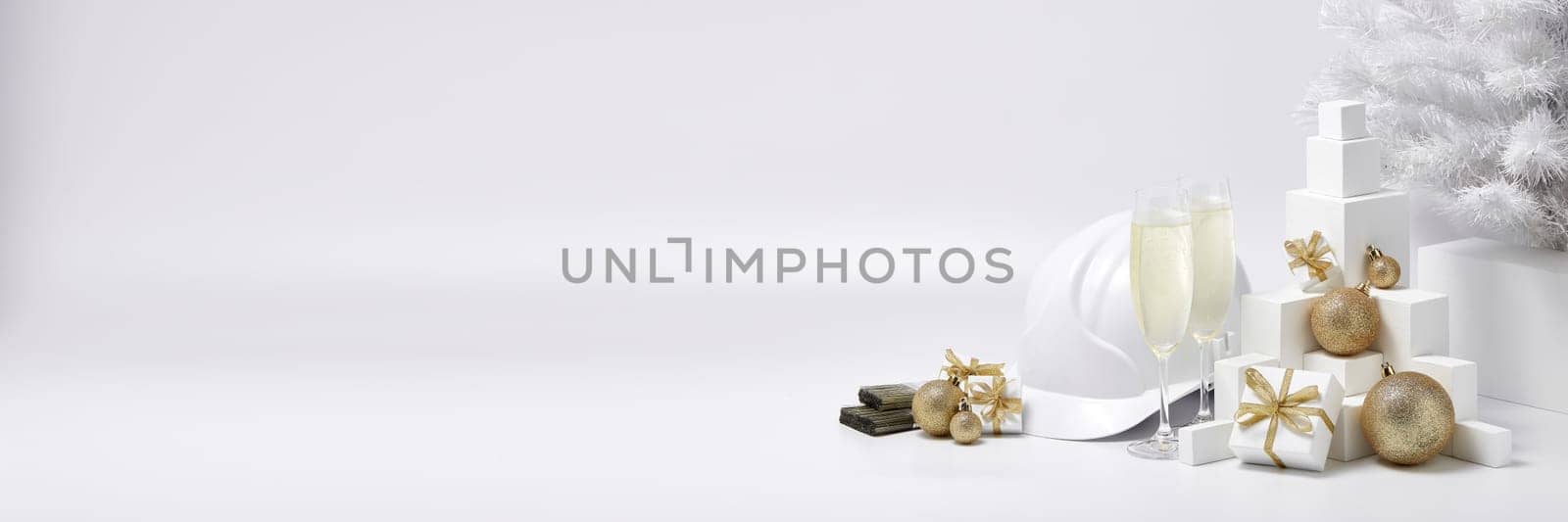 Construction hard hat, white fir tree, two glasses with champagne, gift boxes and Christmas ornaments on a white background with copy space. New Year and Christmas construction background