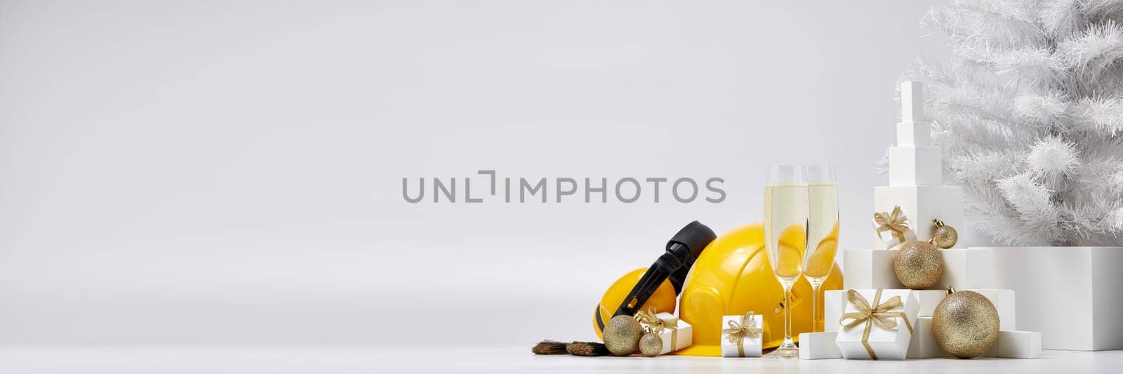 Construction protective earphones and hard hat, fir tree, flutes with champagne, gift boxes and Christmas ornament on a white background with copy space. New Year and Christmas construction