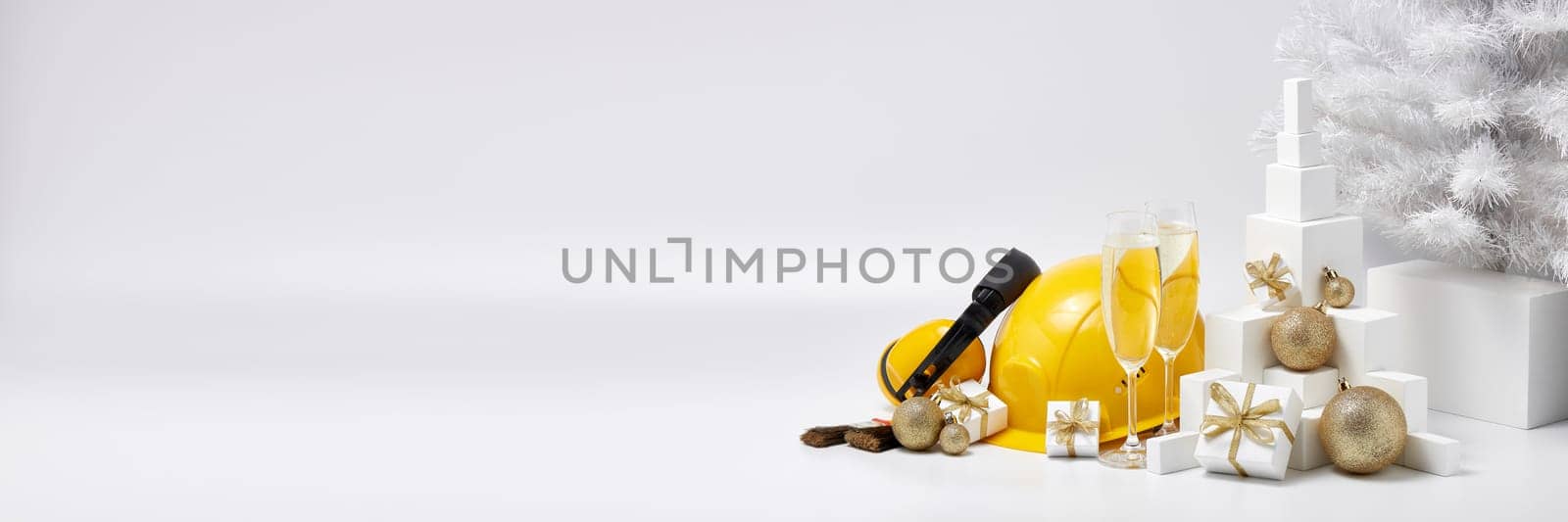 Construction protective earphones and hard hat, fir tree, flutes with champagne, gift boxes and Christmas ornament on a white background with copy space. New Year and Christmas construction