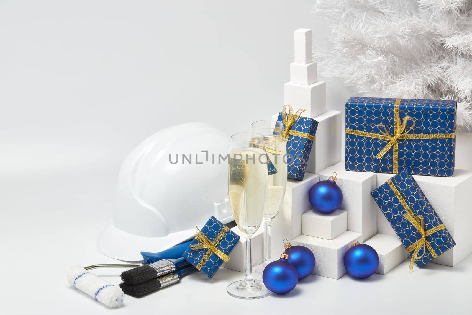 Construction hard hat, white fir tree, two glasses with champagne, gift boxes and Christmas ornaments on a white background. New Year and Christmas construction background