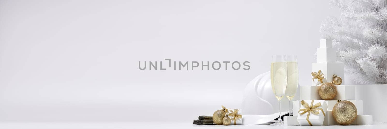 Construction hard hat, white fir tree, two glasses with champagne, gift boxes and Christmas ornaments on a white background with copy space. New Year and Christmas construction background