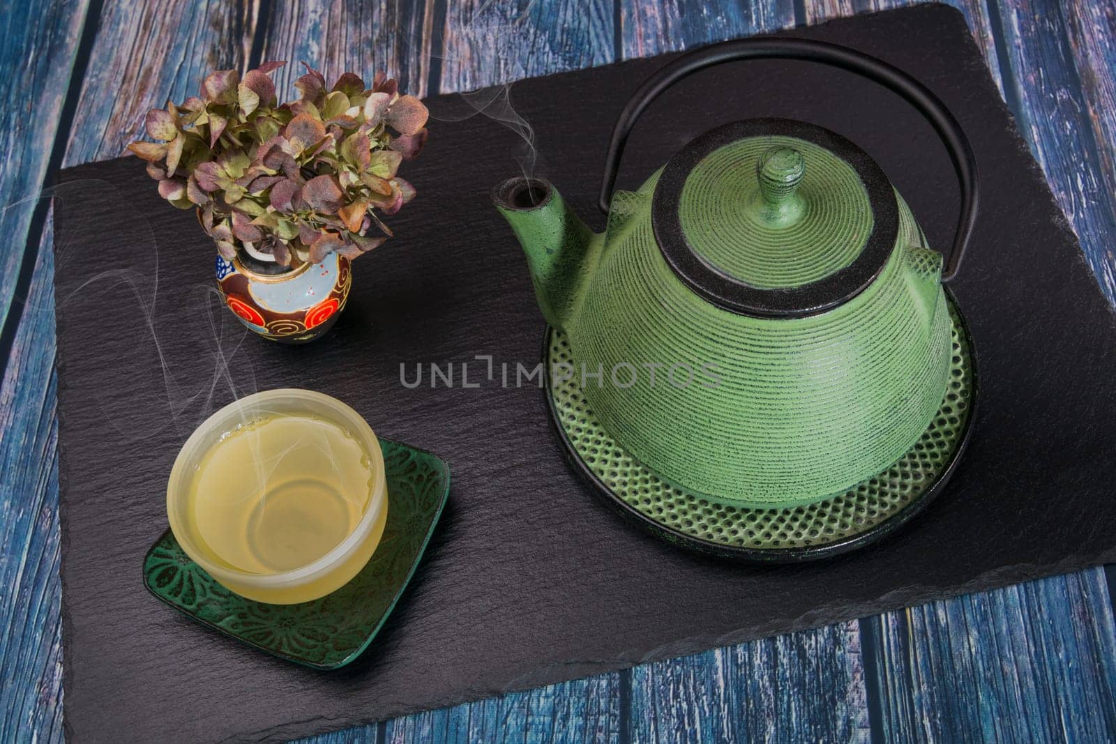 A steaming green metal teapot, a glass bowl with steaming tea and a chinese vase with dried hydrangea on a slate slab.