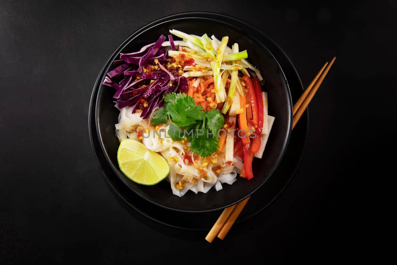 Spicy rice noodel salad in a black bowl with chopsticks, including peppers, cabbage, scallions, and chili lime dressing