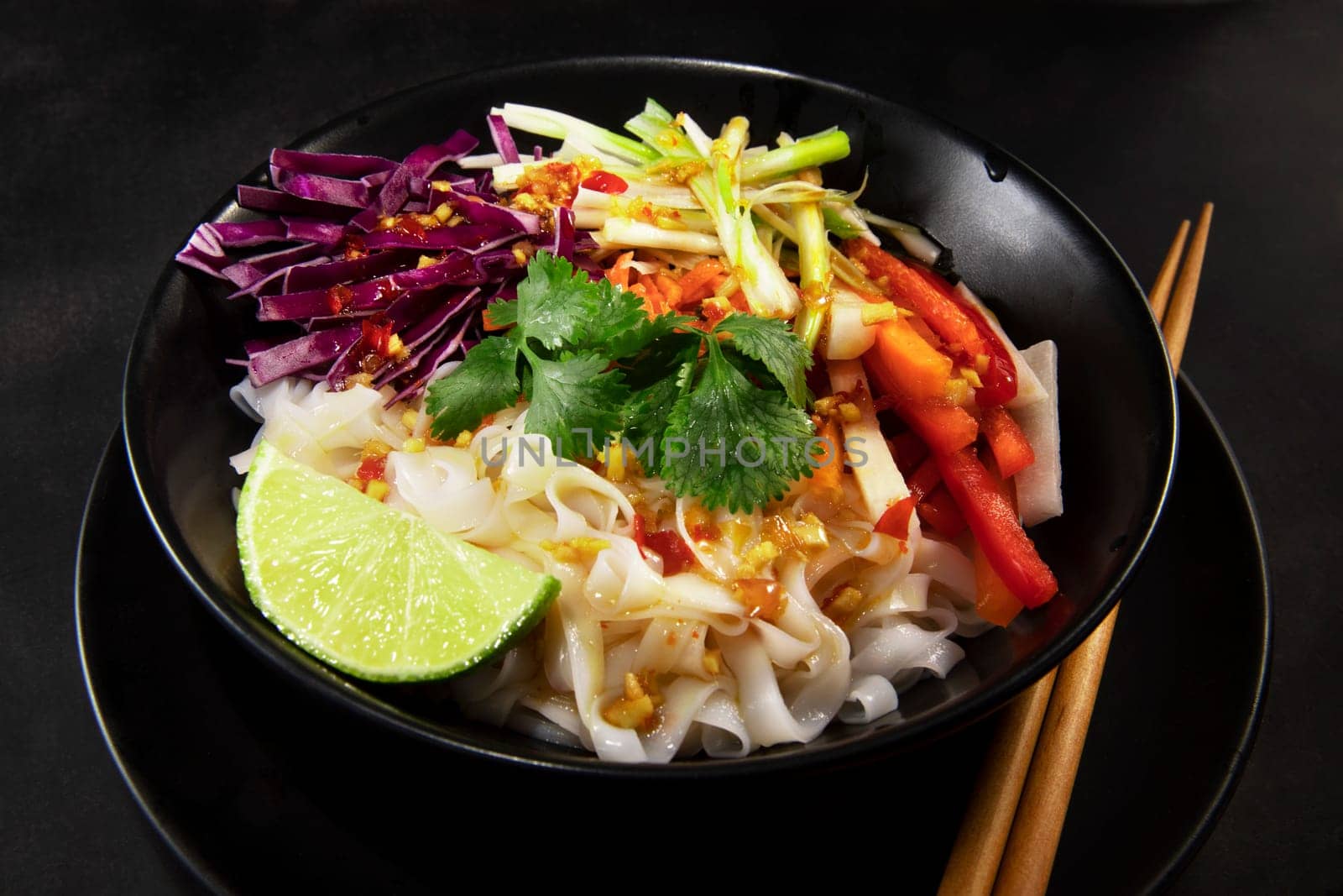 Spicy rice noodel salad in a black bowl with chopsticks, including peppers, cabbage, scallions, and chili lime dressing