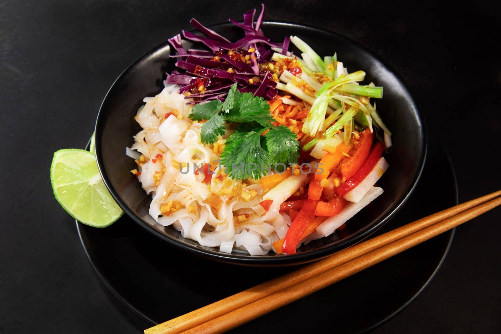 Spicy rice noodel salad in a black bowl with chopsticks, including peppers, cabbage, scallions, and chili lime dressing