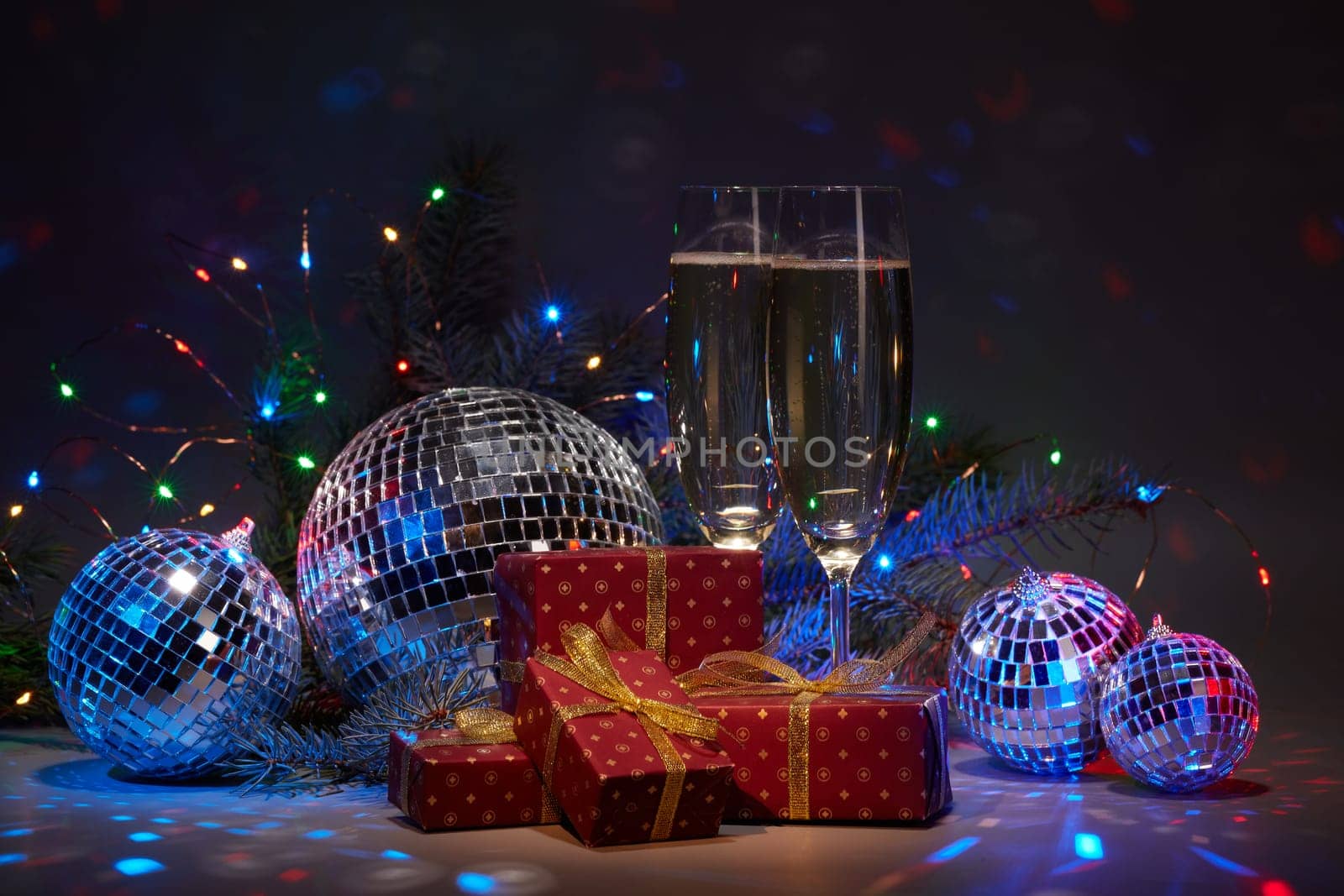 Champagne flutes, mirror balls (disco ball), fir branch, on dark background with colorful LED lights garland. New Year and Christmas. Weekend or holiday party.