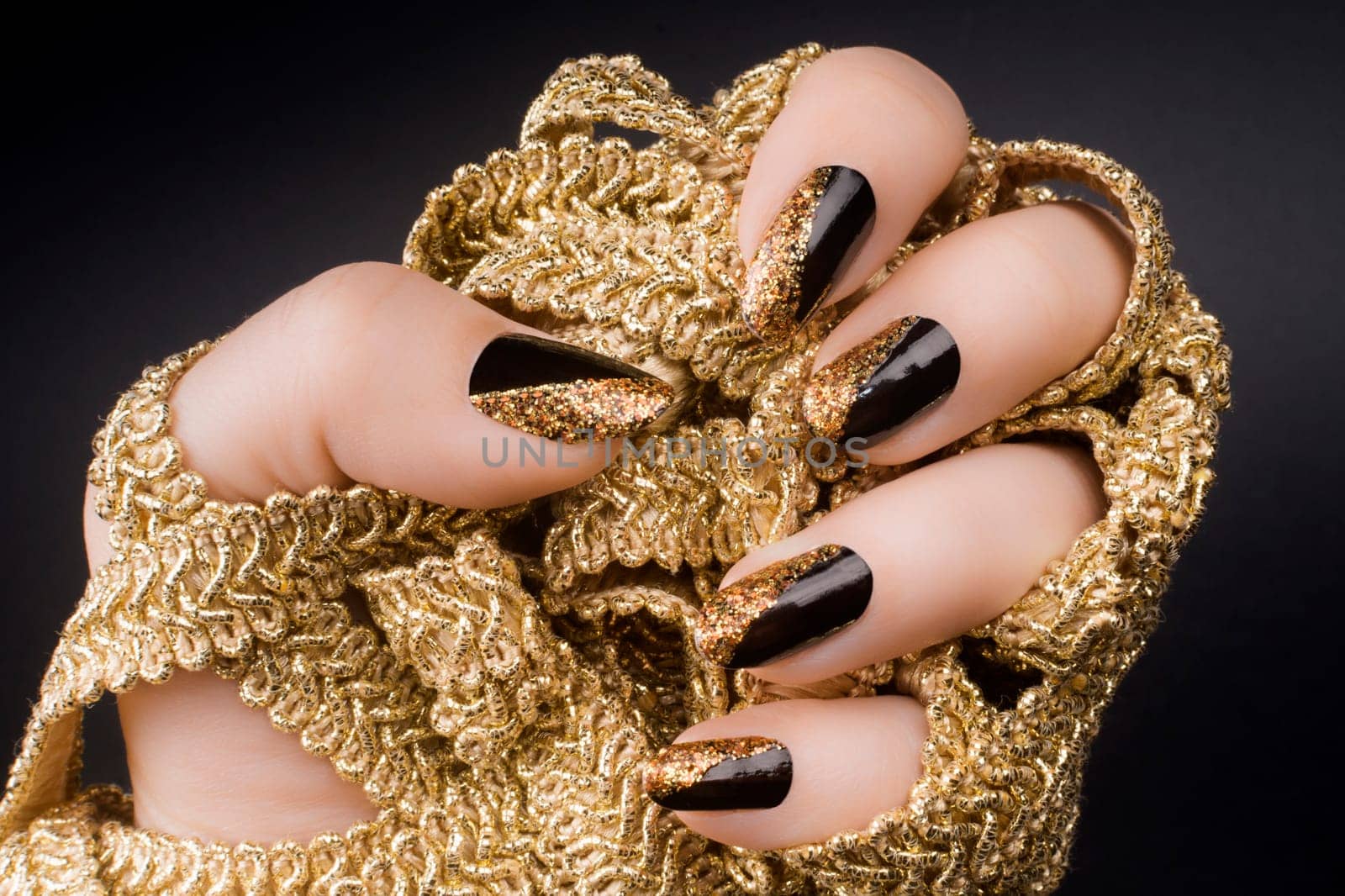 Female hand with golden black nails is holding a golden decoration on black background.