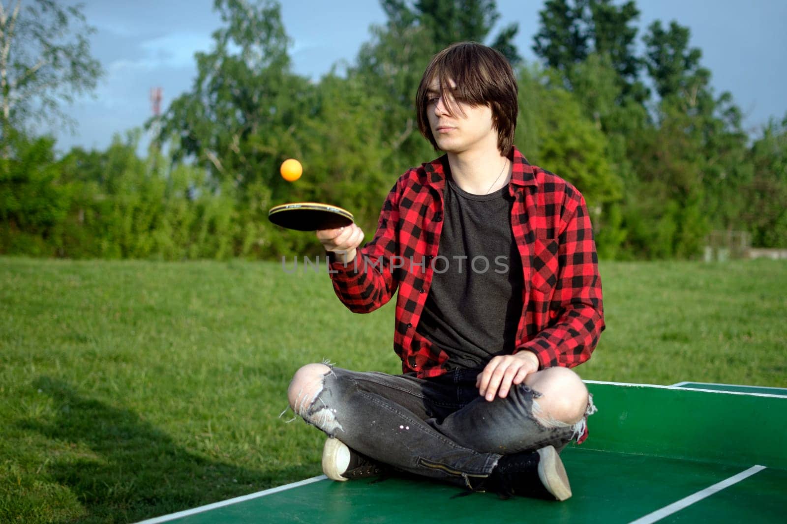 The teen boy is sitting on the green table for table tennis and playing alone with racket and ball outdoors.
