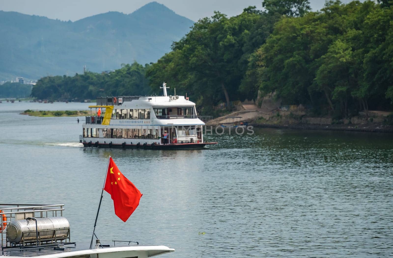 Guilin, China - August 2019 : Red Chinese national  flag fluttering on a mast on the deck of sightseeing boatt full of tourists departing on a trip on the magnificent Li river from Guilin to Yangshuo