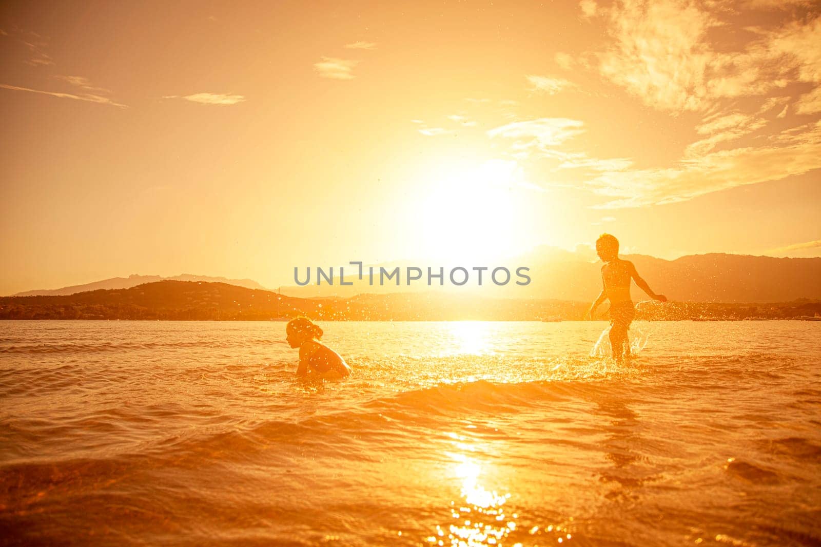 pair of children playing in the sea, shot taken at sunset, hills in the background. Corsica.