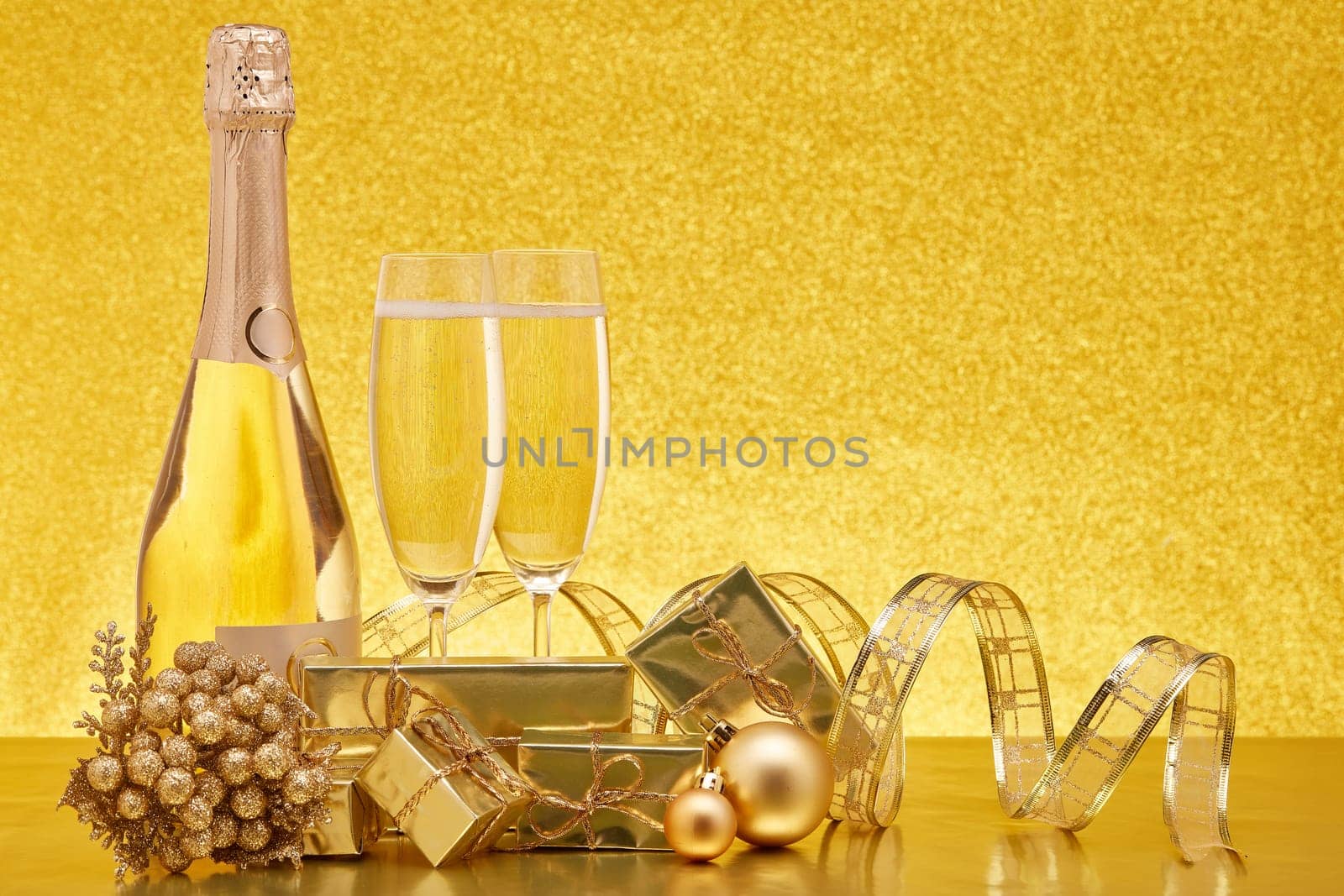 Golden Christmas ornaments, gift boxes, champagne bottle and full champagne flutes on gold background with copy space. Christmas. New Year. Design element for greeting card, invitation, advertising