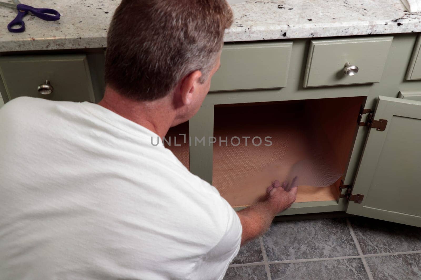 In a home bathroom inside a green painted wood cabinet a man lines the bottom of a bathroom cabinet with a plastic sheet. Clear plastic shelf liner being put on the bottom of a home bathroom cabinet.