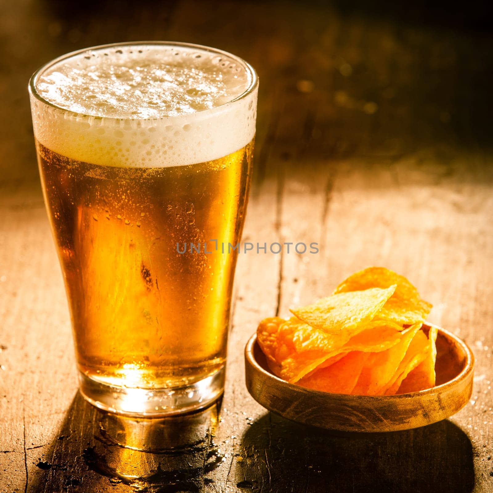 Glass of refreshing cold beer with a small bowl of potato crisps served on a rustic wooden bar counter , square format with copyspace