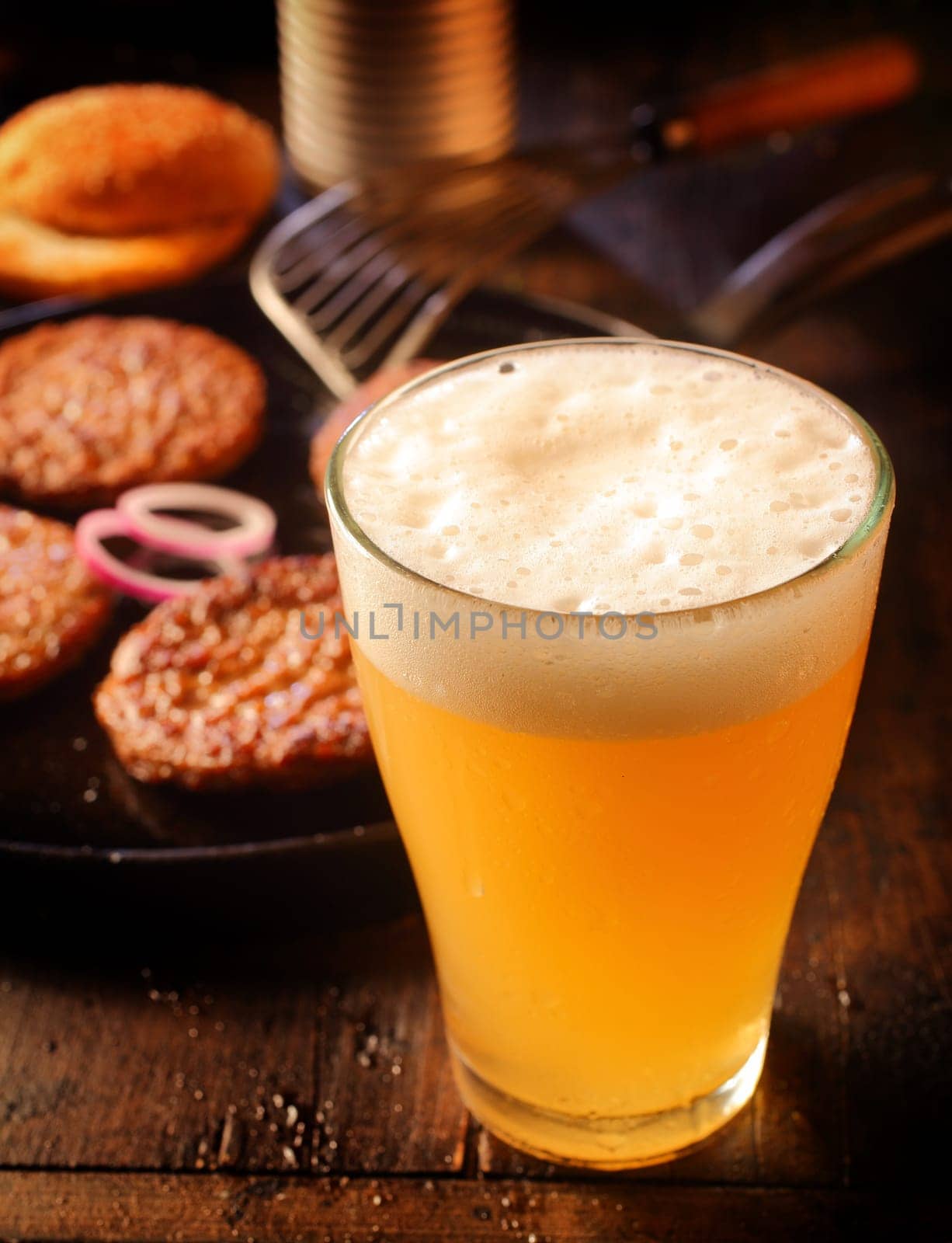 Cold glass of frothy beer standing alongside a pan of grilled beef burger patties in a pub or restaurant , high angle with focus to the frothy head of the lager