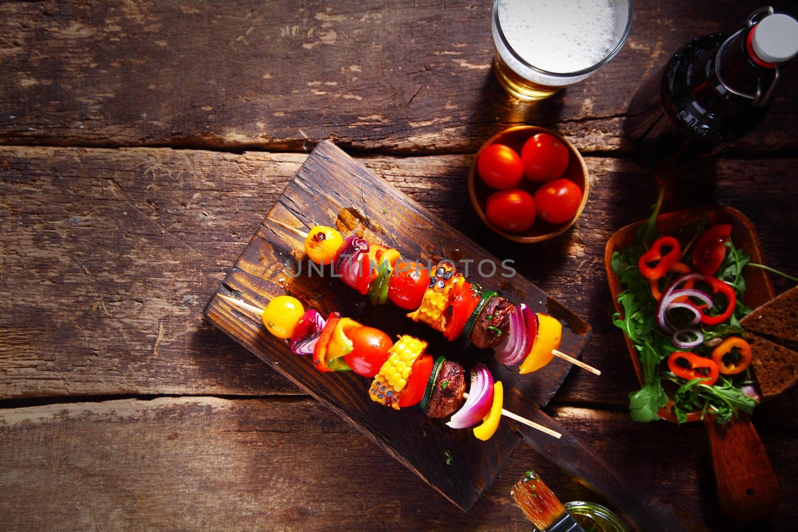 Delicious bar lunch of fresh vegan or vegetarian kebabs with assorted roasted colorful vegetables on skewers served on a rustic wood table with a beer and salad, with copyspace