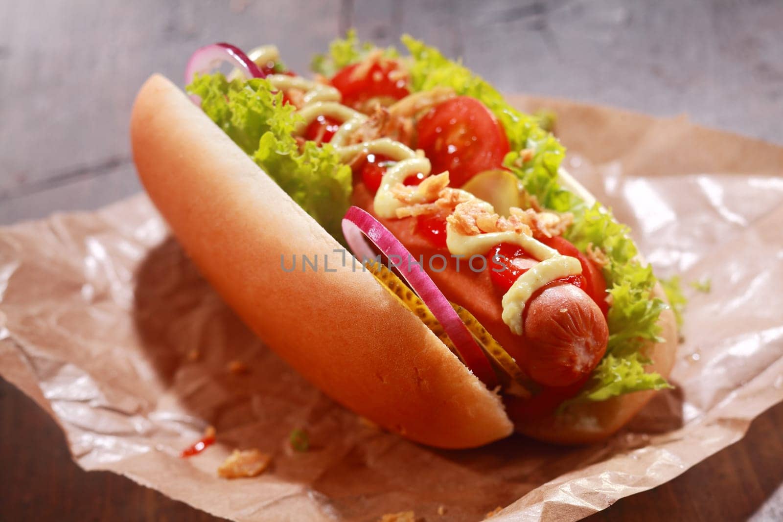 Tasty homemade hot dog with sausage garnished with green salad or lettuce, onion, pickles, tomatoes and mustard, served on a paper