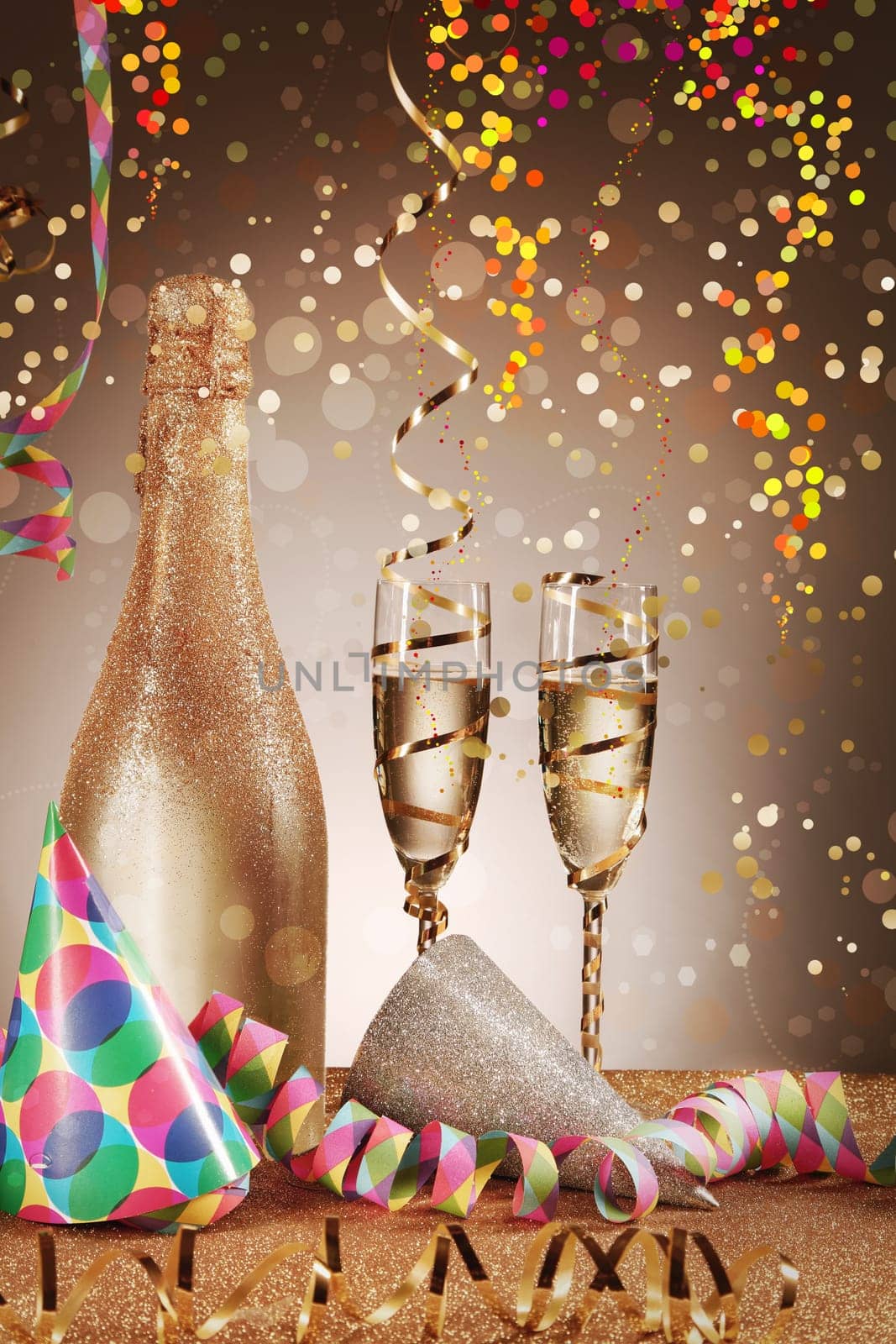 Celebration Concept - Close up Cone Hats and Champagne Bottle and Glass on Glittery Table with Paper Streamers. Emphasizing Confetti Effect on Gradient Brown.
