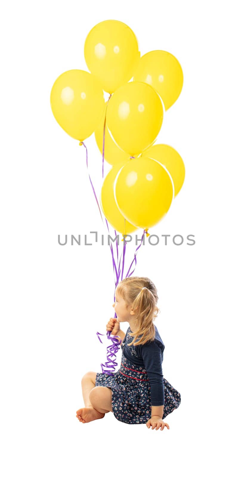 portrait of a sitting 3 year old girl holding a group of yellow balloons. side view. isolated on white