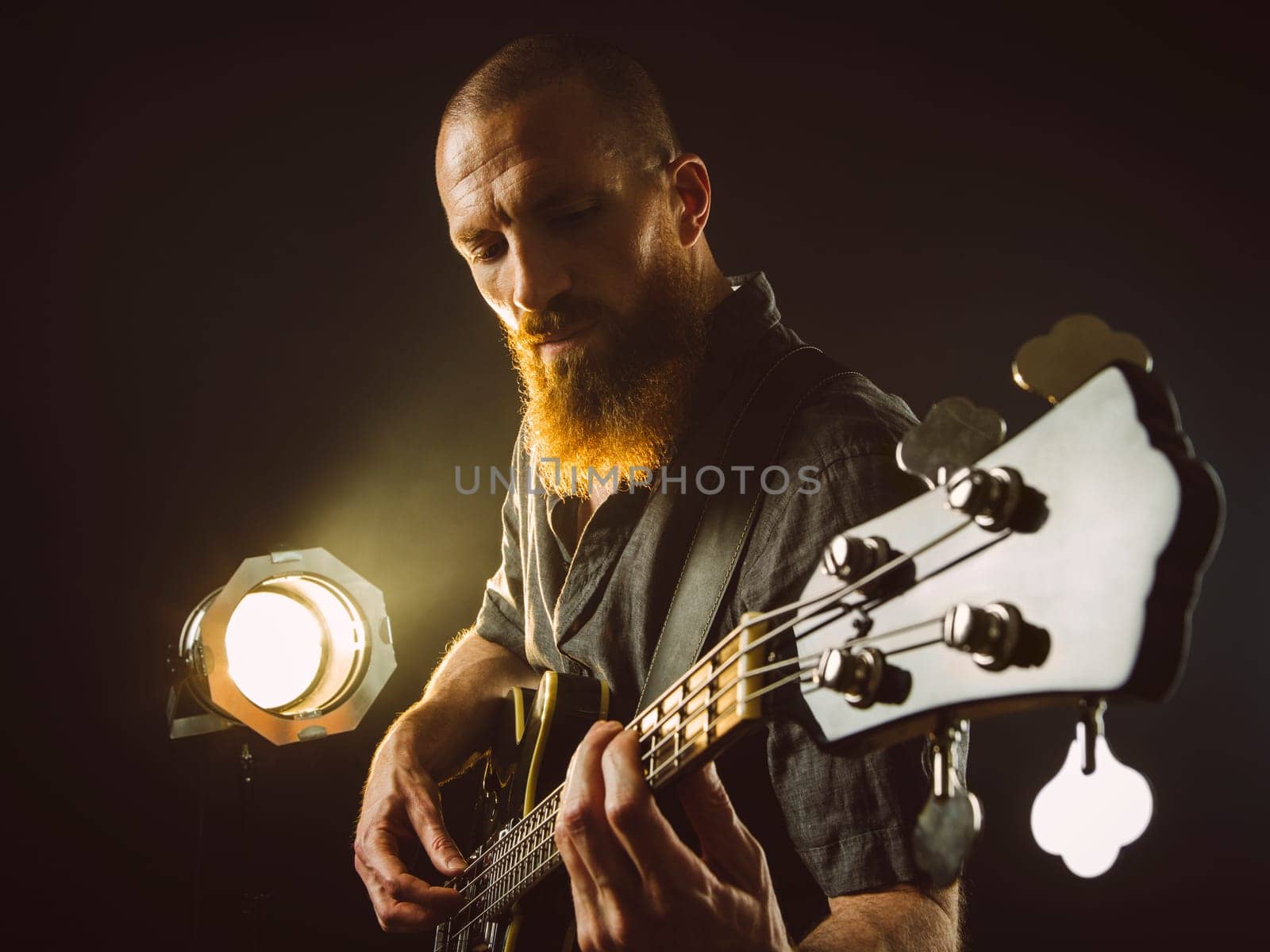 Photo of a bearded man playing bass guitar on stage in front of spotlights..