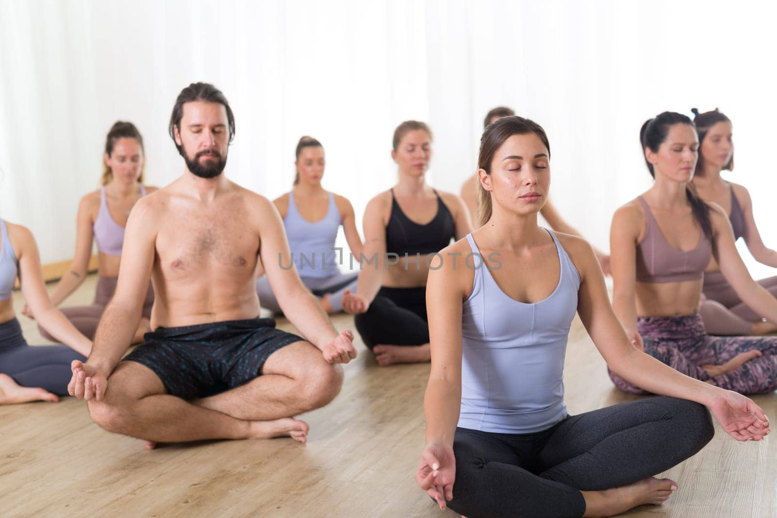 Group of young sporty attractive people in yoga studio, practicing yoga lesson with instructor, sitting on floor in Siddhasana, easy seated yoga pose. Healthy active lifestyle, working out in gym.