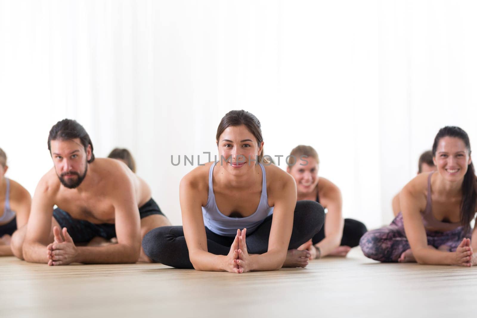 Group of young sporty attractive people in yoga studio, practicing yoga lesson with instructor, sitting on floor in forward band stretching yoga pose. Healthy active lifestyle, working out in gym.
