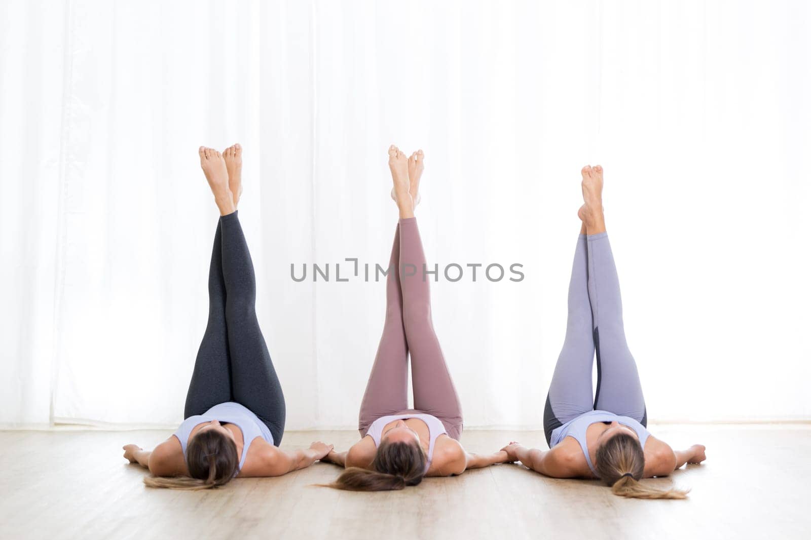 Group of three young sporty attractive women in yoga studio, lying on the floor, holding hands, legs up against white background. Healthy active lifestyle, working out indoors in gym.