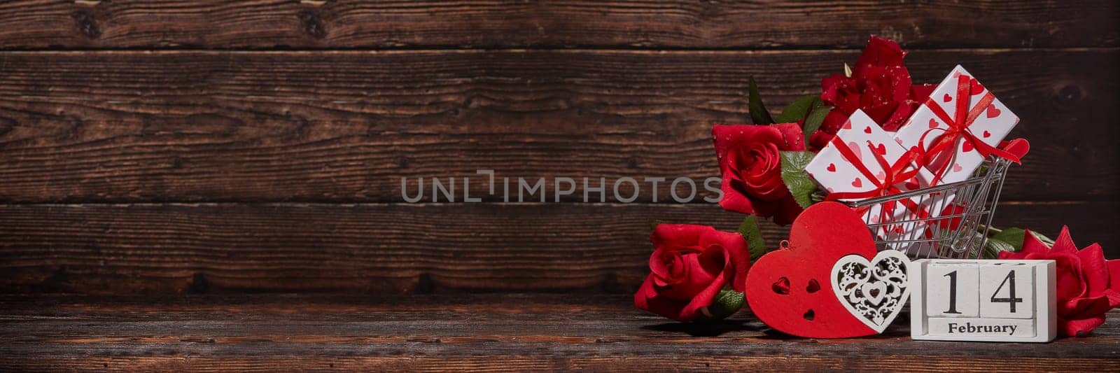Shopping trolley, gift boxes, calendar with date 14 february, red roses and hearts on brown wooden background with copy space. Valentine's Day. Extra wide panorama banner background