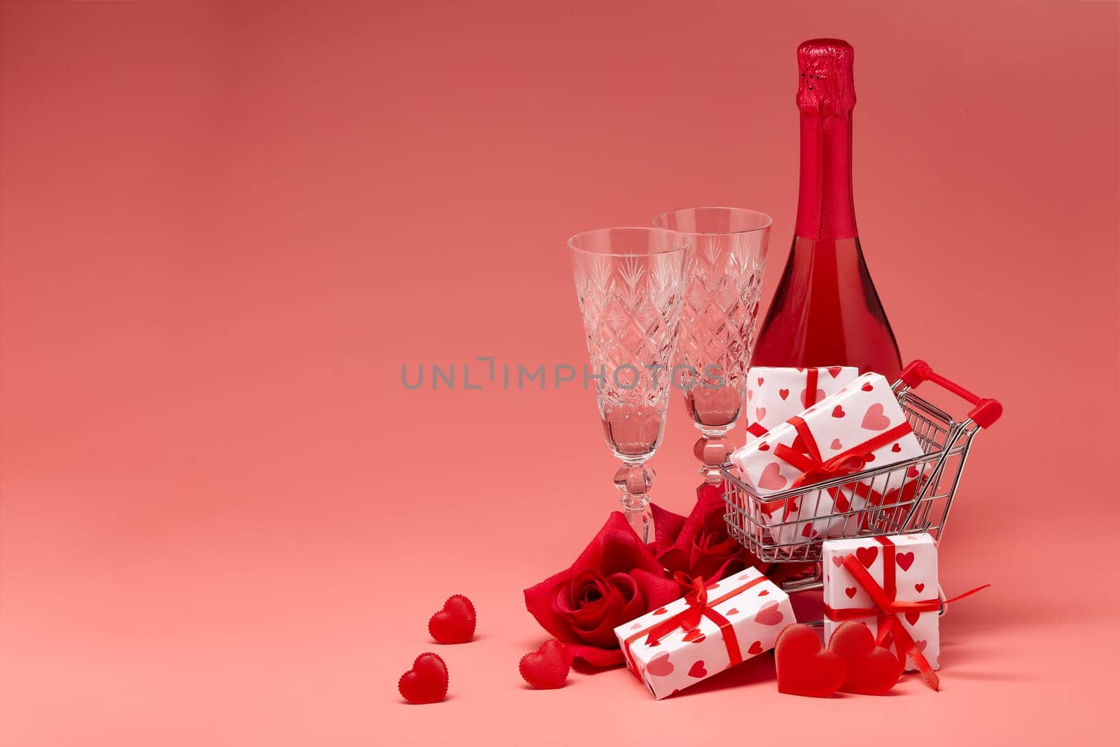 Shopping trolley, gift boxes, red roses and hearts, champagne and flutes on pink background with copy space. Valentine's Day. Festive background with decorative hearts.Valentine's Day. Wedding day.