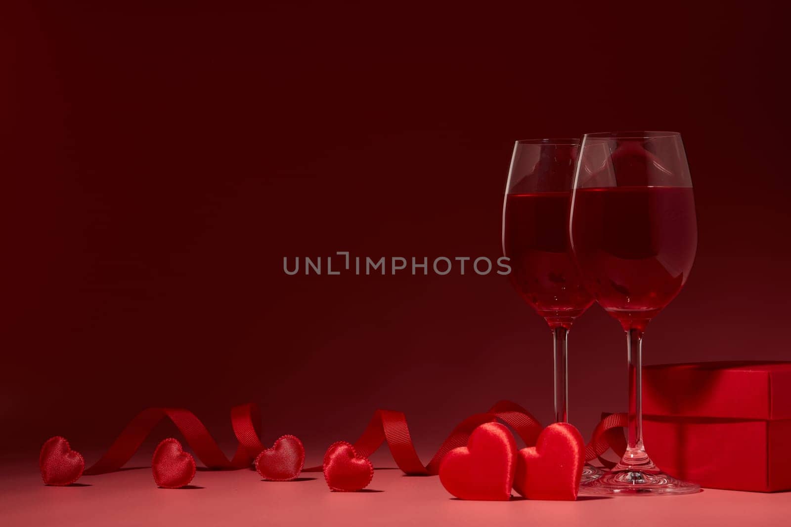 Gift box, red hearts, two glasses with wine on red background. Valentine's Day card. Copy space. Design element for romantic greeting card, wedding invitation.