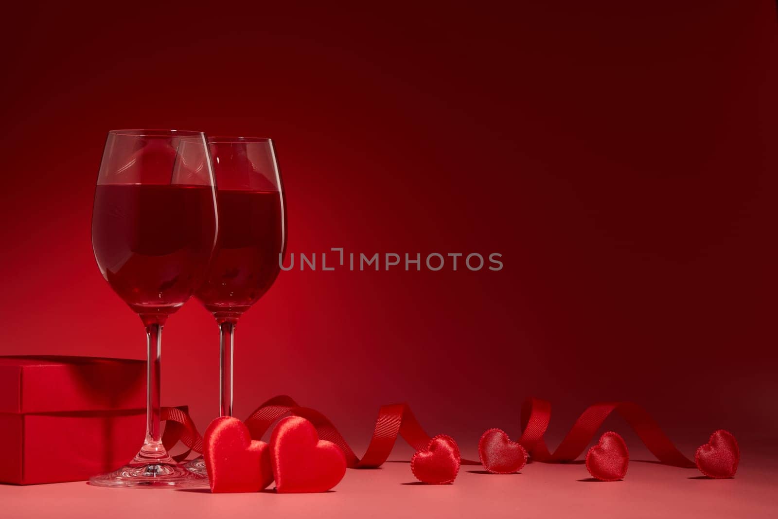 Gift box, red hearts, two glasses with wine on red background. Valentine's Day card. Copy space. Design element for romantic greeting card, wedding invitation.