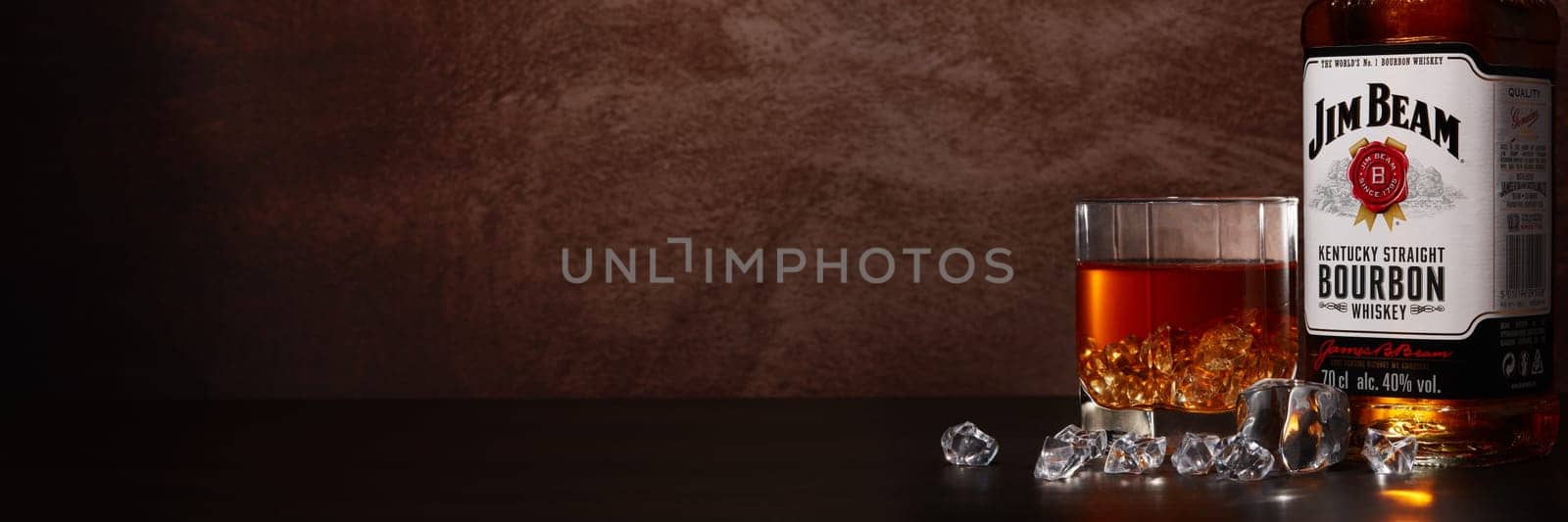 St.Petersburg, Russia - December 2019 - Bottle of Jim Beam bourbon whiskey and glass with drink and ice on wooden table on brown background with copy space.  Extra wide panorama banner background