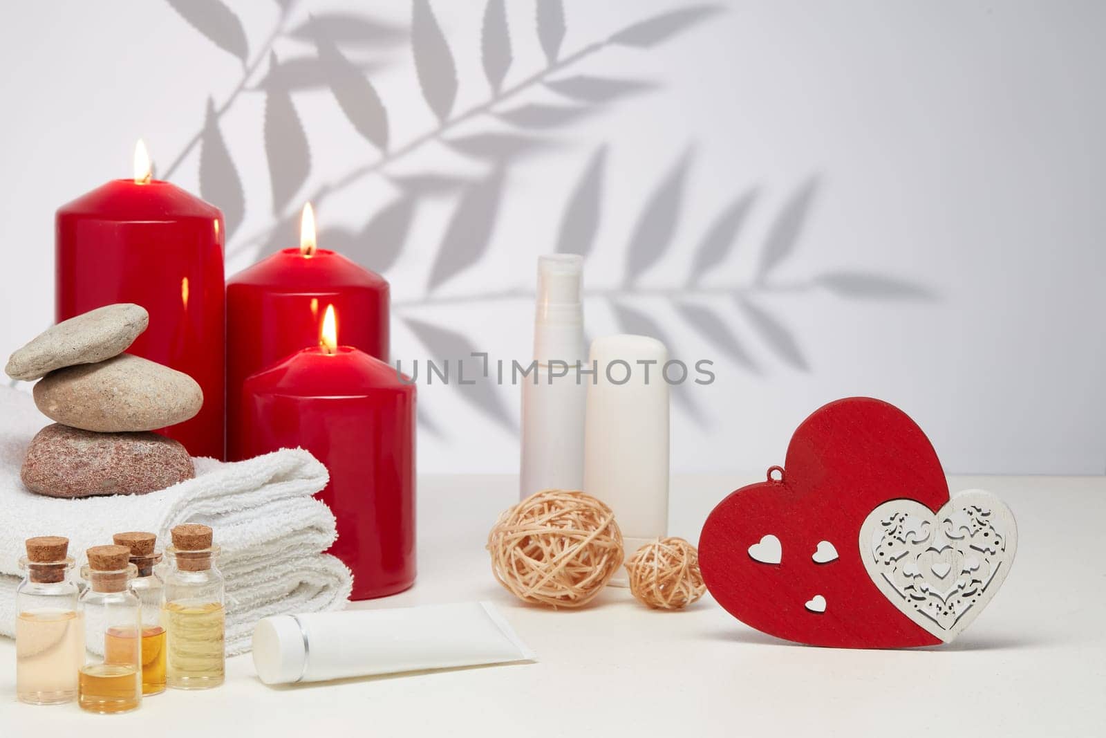 Spa still life with creams, essential oils, burning red candles, hearts on light background with shadow from plant. Healthy lifestyle, body care, Spa treatment. Valentine's Day. Honeymoon. Wedding